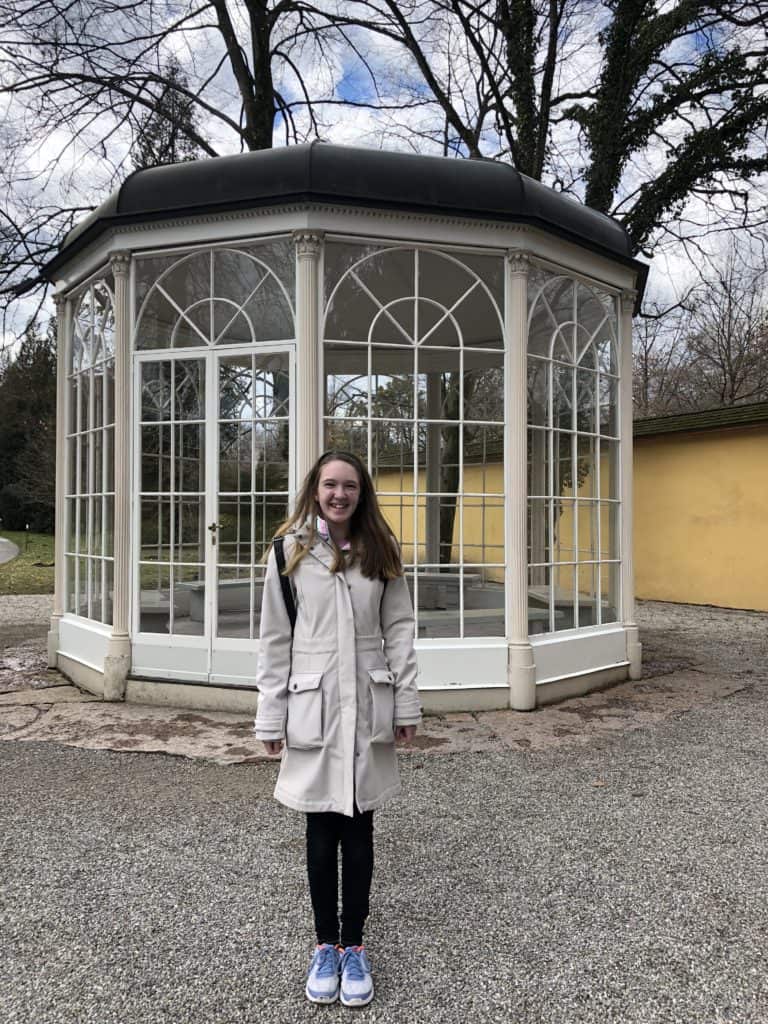 Girl standing outside of white gazebo with dark roof and glass windows from The Sound of Music.