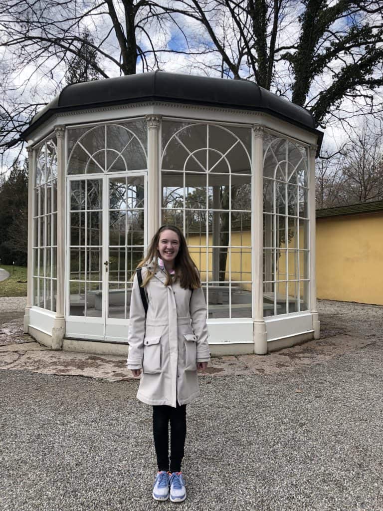 Girl standing outside of white gazebo with dark roof and glass windows from The Sound of Music.