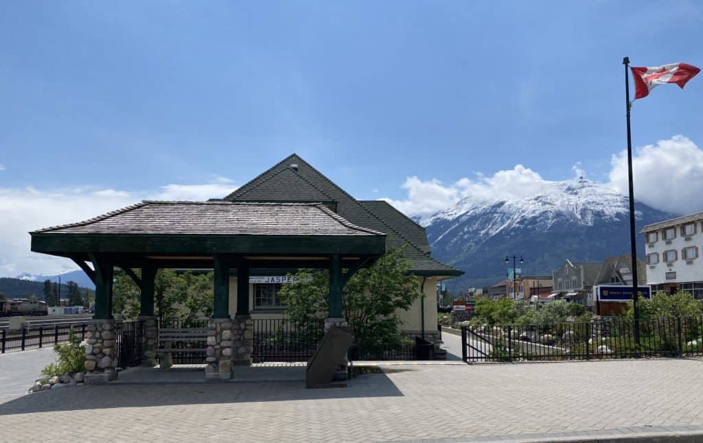 Canadian flag beside Jasper train station with mountains in background.