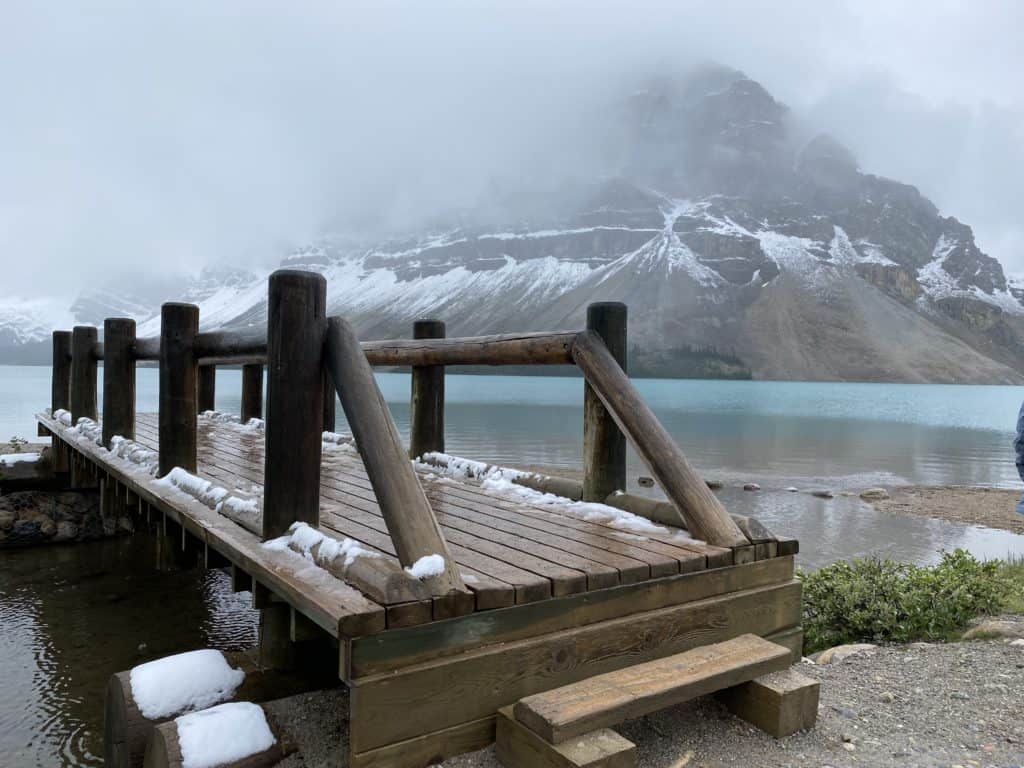 Wooden bridge with snow on it at Bow Lake on foggy day with snow topped mountain in background.