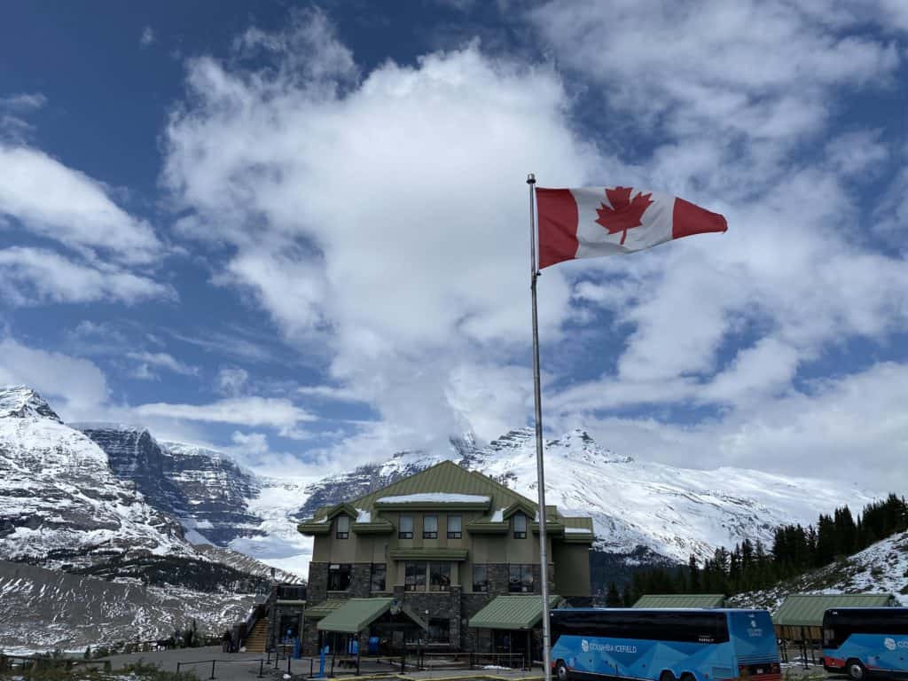 Blue sky with fluffy white clouds and Canadian flag flying outside the Glacier Discovery Centre and Glacier View Lodge with snow-covered mountains in background.