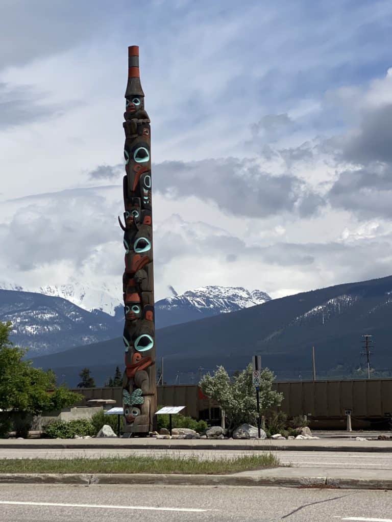 Totem pole in Jasper with cloudy sky and snow-topped mountains in background.