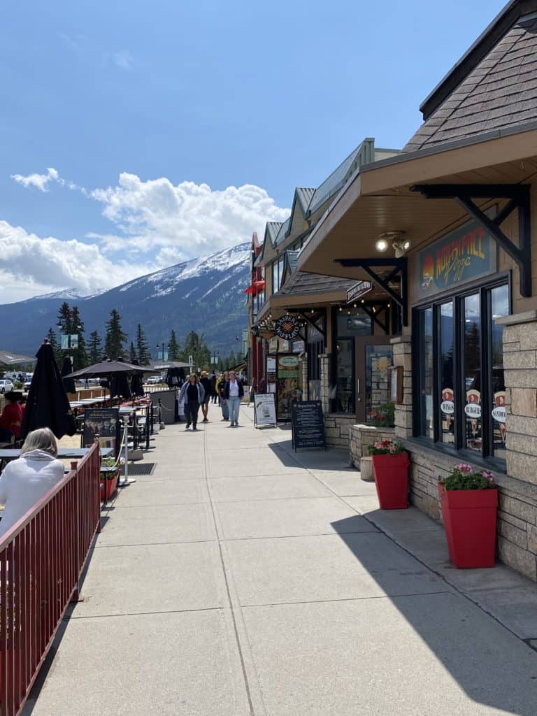 People walking down sidewalk in Jasper with shops on right, outdoor dining on the left and mountains in the background.