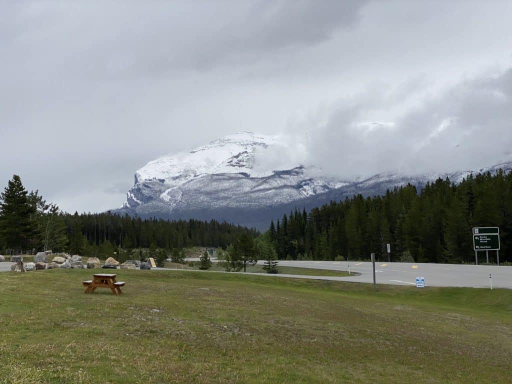 View of snow-covered mountains at Saskatchewan River Crossing alongside highway with picnic table on lawn in foreground.