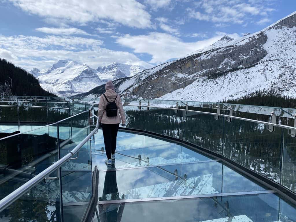 Young woman in pink coat and hat walking on glass skywalk with mountains around.