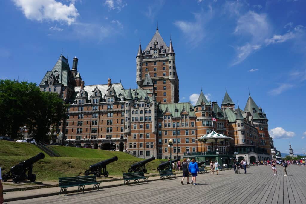 People walking along boardwalk in front of Chateau Frontenac on a summer day with blue sky and a few white cloud.