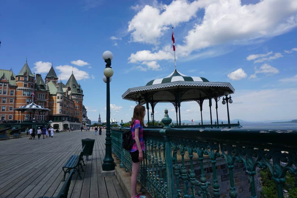 Girl in black shorts and pink striped shirt standing beside green railing looking out at St. Lawrence River from Dufferin Terrace with Chateau Frontenac in background.
