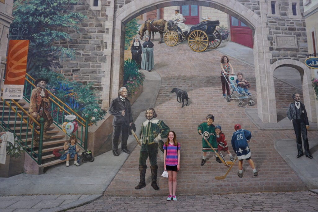 Young girl in pink and purple striped shirt standing in front of mural in Quebec City.