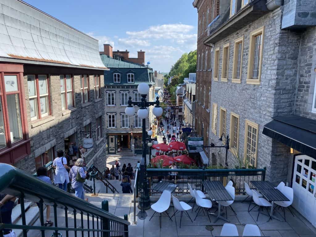 View down the stairs to Rue Petit Champlain in Quebec City - people walking down stairs and along street between grey stone buildings with colourful window frames.