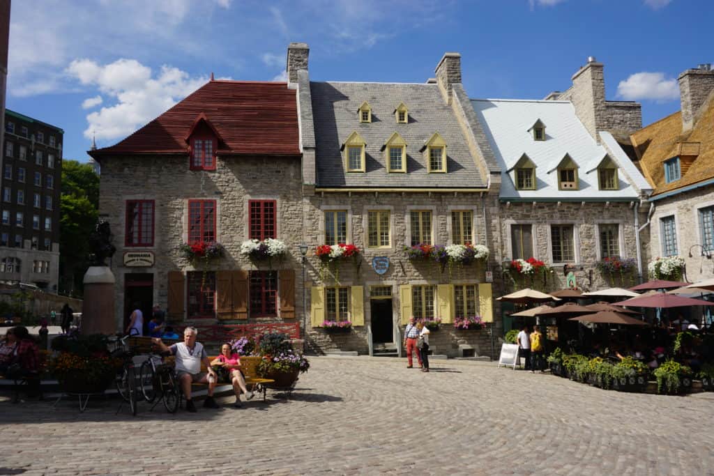Historic stone buildings with flowers in window boxes in Place Royal in Quebec City with people standing and sitting in square.