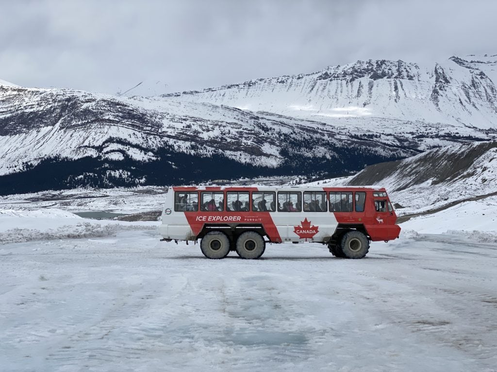 Large red and white Ice Explorer all-terrain vehicle parked on the Athabasca Glacier with snow covered mountains in background.