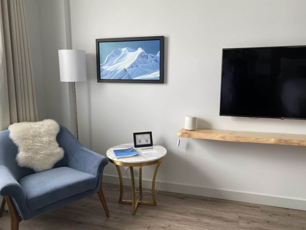 Seating area in room at Glacier View Lodge - blue upholstered chair with white furry throw, floor lamp, picture of snow covered mountain haning on wall, small side table with metal base and white top, tv on wall with wooden shelf under.