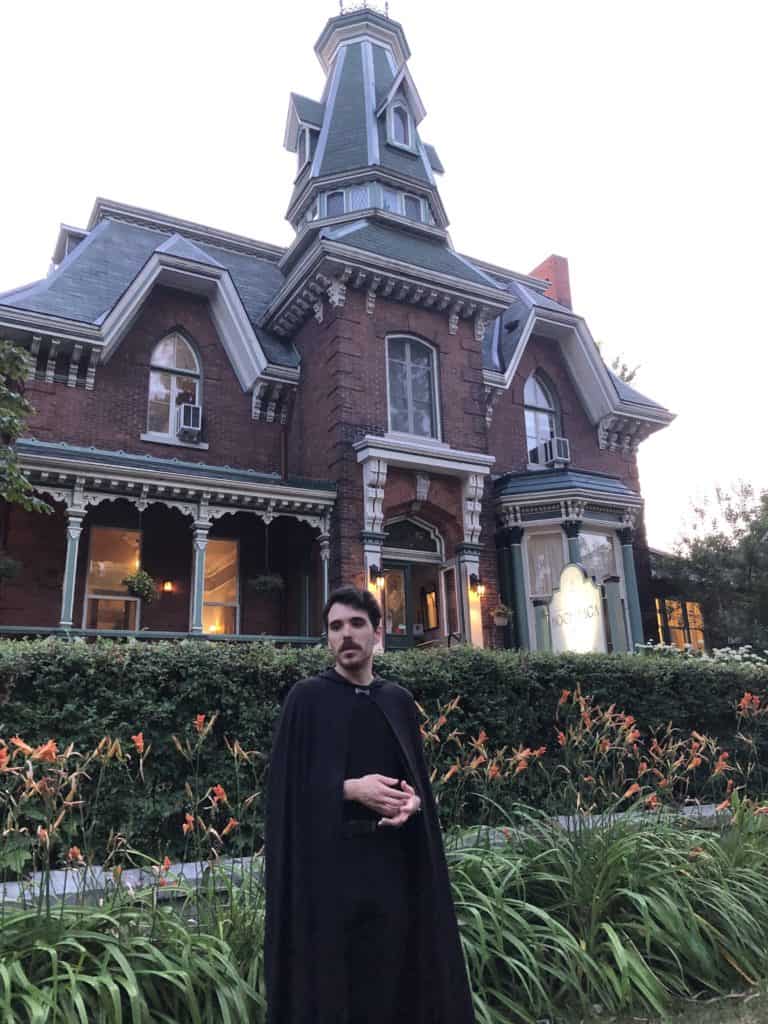 Man wearing black pants, shirt and a black cape standing in front of Hochelaga Inn in Kingston, Ontario at dusk.