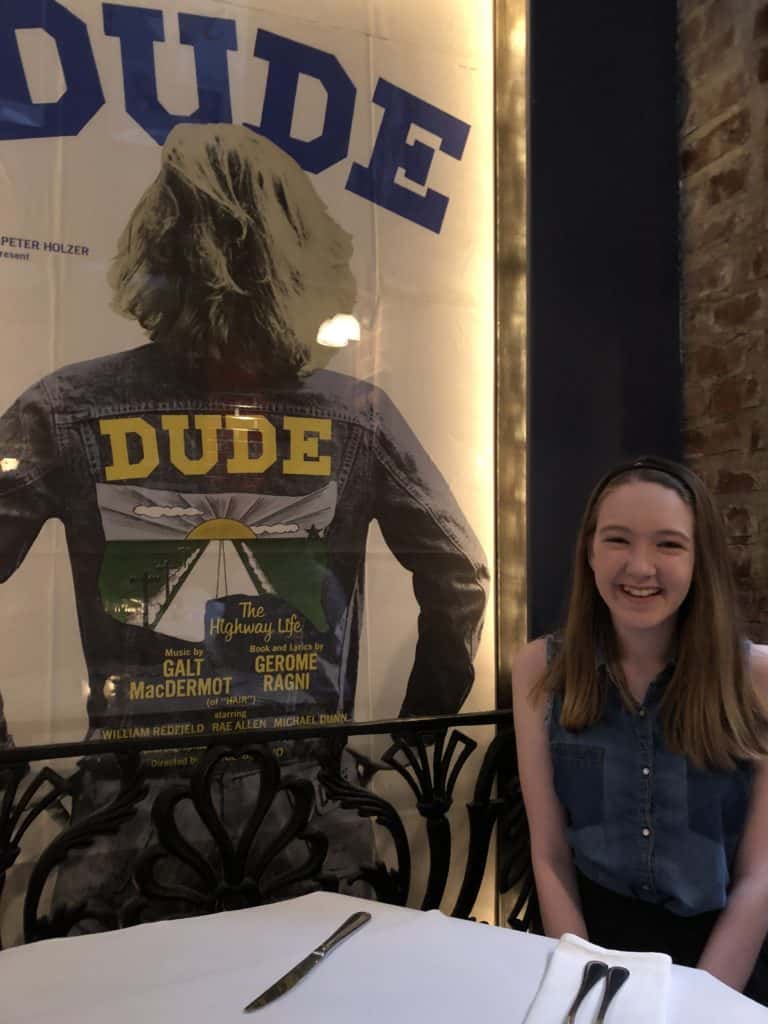 Teen girl sitting at table beside poster for Dude at Joe Allen Restaurant in New York City.