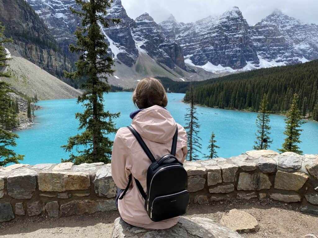 Young woman wearing pink jacket and small black backpack sitting in front of rock wall looking out at bright blue Moraine Lake.