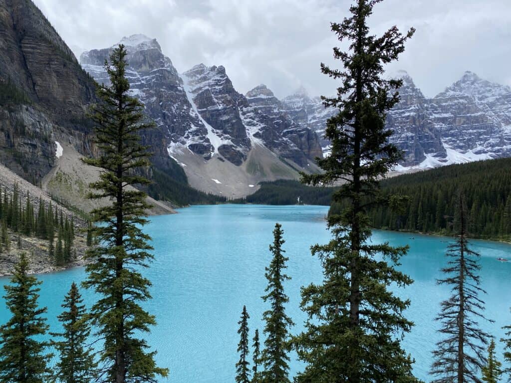 Moraine Lake, Alberta - turquoise blue lake with evergreen trees in foreground and snow-capped mountain peaks in background.