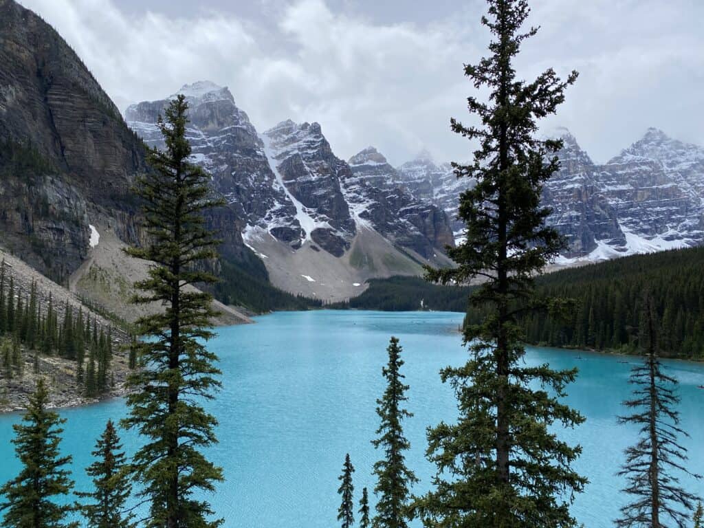 Bright blue Moraine Lake with evergreen trees in front and snow-capped mountains in background.