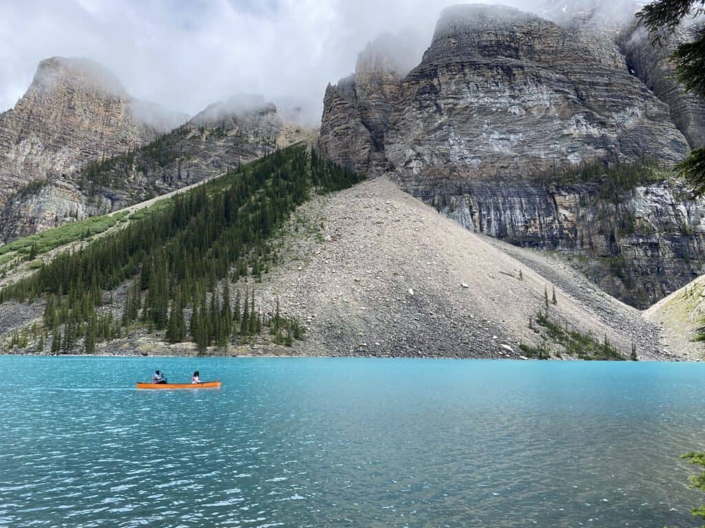 Two people paddling an orange canoe on bright blue waters of Moraine Lake, Alberta with mountains in background.