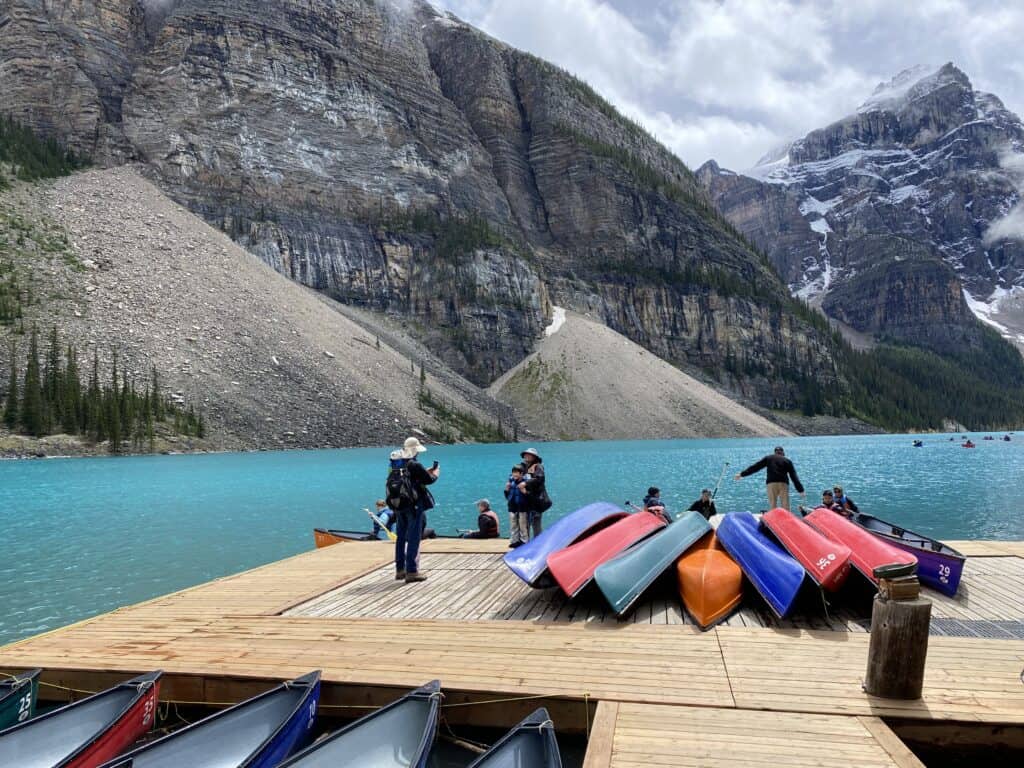 People standing on wooden dock with several brightly coloured canoes - bright blue Moraine Lake and mountains in background.