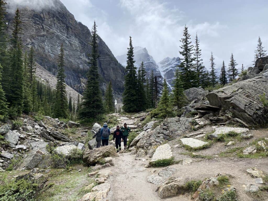 People walking rocky path with trees and mountains in background.