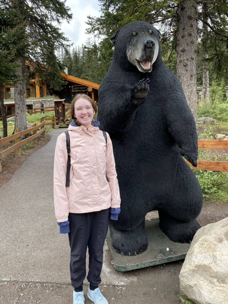 Young woman in pink coat standing with bear sculpture outside Moraine Lake gift shop.