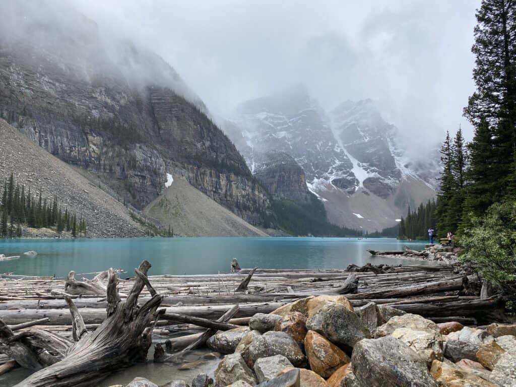 Rocks and driftwood on shores of bright blue Moraine Lake with fog and snow-capped mountains in background.