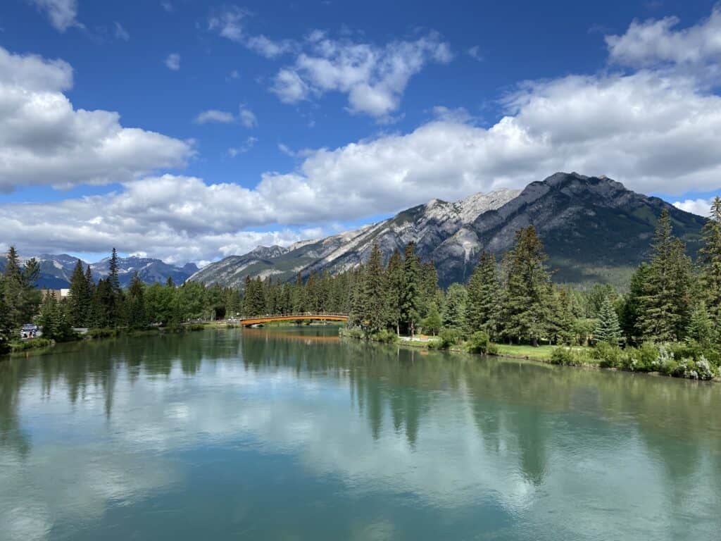Bow River in Banff with bridge and mountains in background on a early summer day with blue sky and fluffy white clouds.