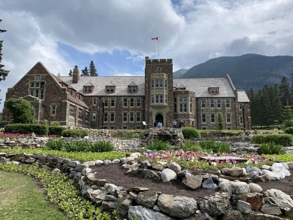 Stone building set amidst gardens with blooming flowers and mountains in the background.