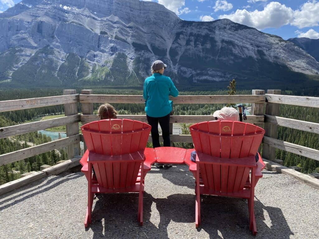 Two women sitting in Parks Canada red chairs and third woman standing in front of them at a railing looking at view of mountains on Banff Hoodoo Trail.