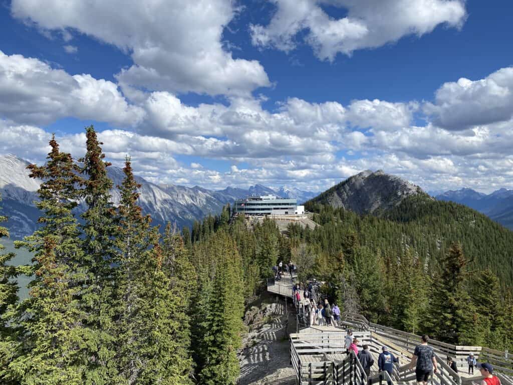 Wooden boardwalk trail at top of Sulphur Mountain with Banff Gondola Terminal and mountains in background.