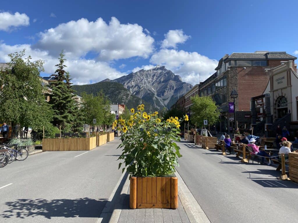 Pedestrian street in downtown Banff - planters of flowers in centre of street, people sitting at outdoor restaurants, mountains in the background.