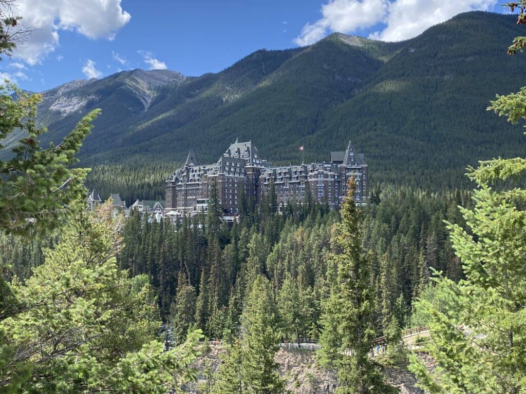 The Fairmont Banff Springs hotel from Surprise Corner viewpoint - surrounded by trees, mountains and blue sky with fluffy white clouds in background.