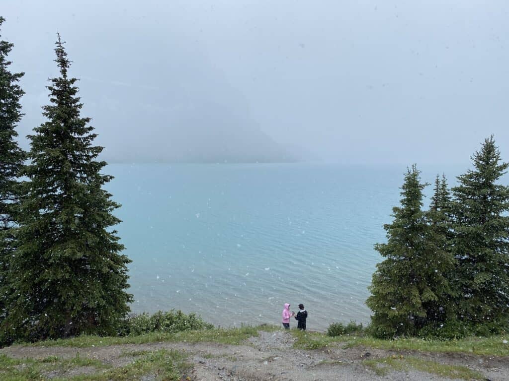 Two people standing between trees with Bow Lake in the background on foggy morning with snow falling.