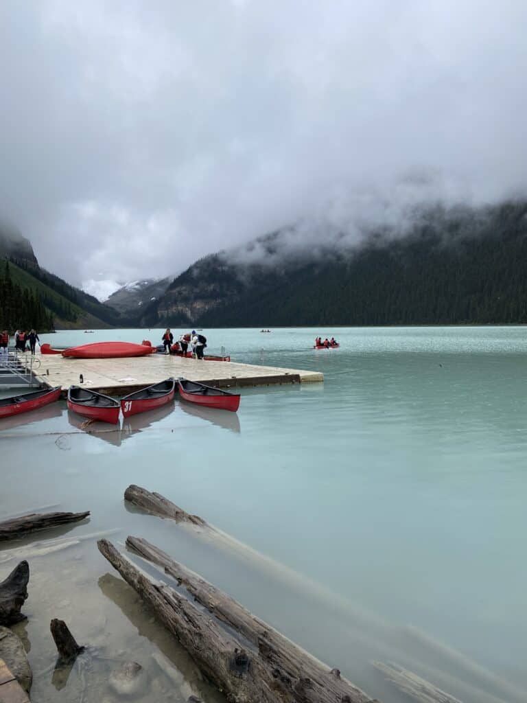 People standing on dock at Lake Louise with bright red canoes on foggy morning with mountains in background.