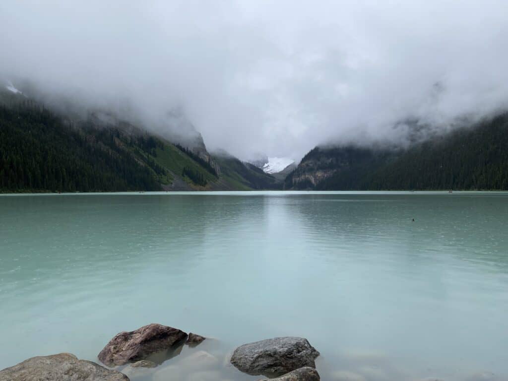 Blue green water of Lake Louise in Banff National Park with low cloud cover over mountains in background.