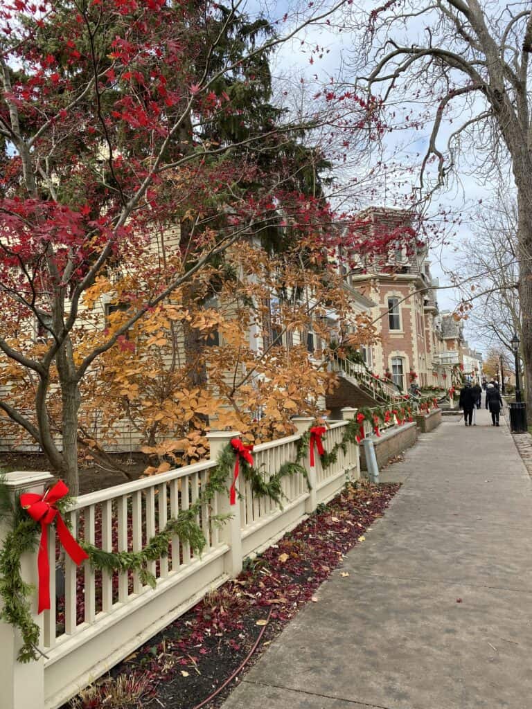 People walking on Queen Street alongside the Prince of Wales Hotel with greenery and red bows on wooden fence.