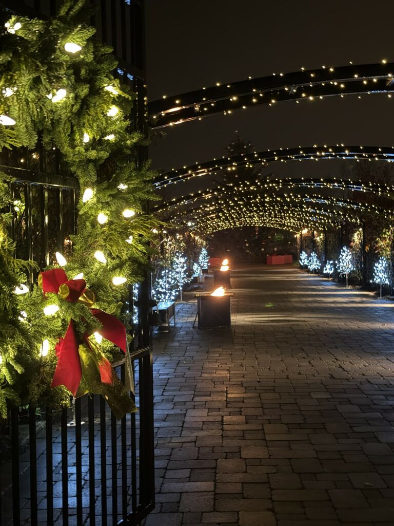Christmas wreath with red bow and white lights mounted on black iron gate with Christmas lights in background at the Christmas Wonderland Garden at Pillar and Post in Niagara-on-the-Lake.