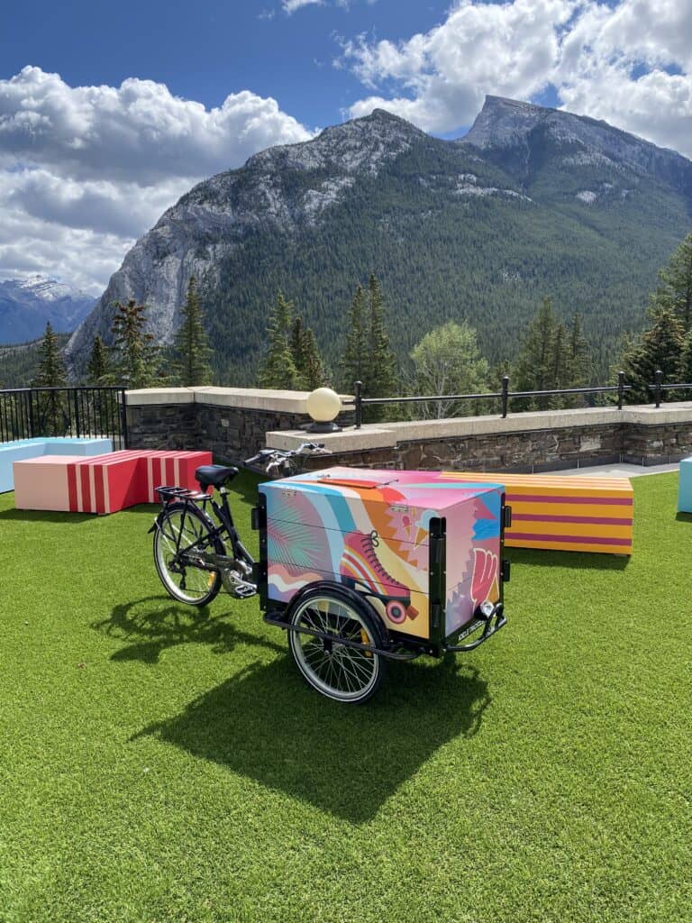 Multi-coloured ice cream cart on bicycle on terrace at Fairmont Banff Springs with trees and mountains in background.