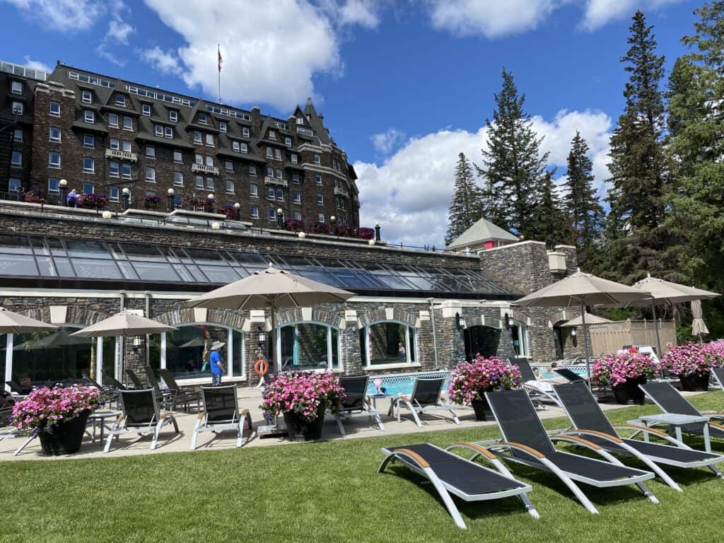 Lounge chairs, planters of bright pink flowers and patio umbrellas around outdoor pool at Fairmont Banff Springs with hotel in background.