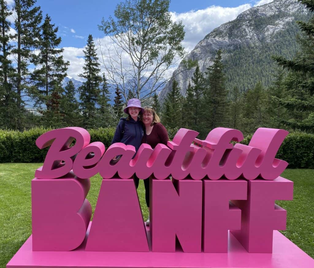 Mom and daughter standing behind dark pink "Beautiful Banff" sign on grounds at Fairmont Banff Springs.
