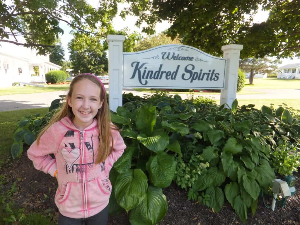 Young girl in pink sweatshirt standing in front of greenery and white and blue sign reading Welcome Kindred Spirits.