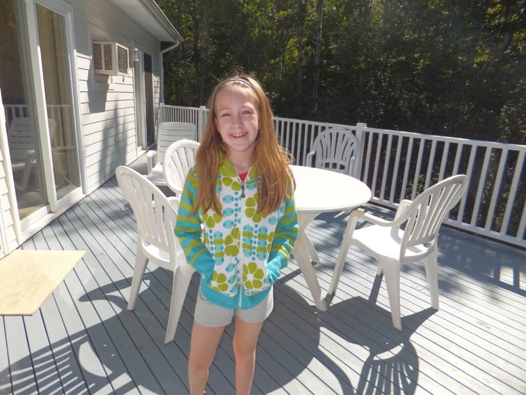 Young girl in grey shorts and blue and green patterned sweatshirt standing on deck in front of white patio table and chairs - Kindred Spirits 3 bedroom cottage.