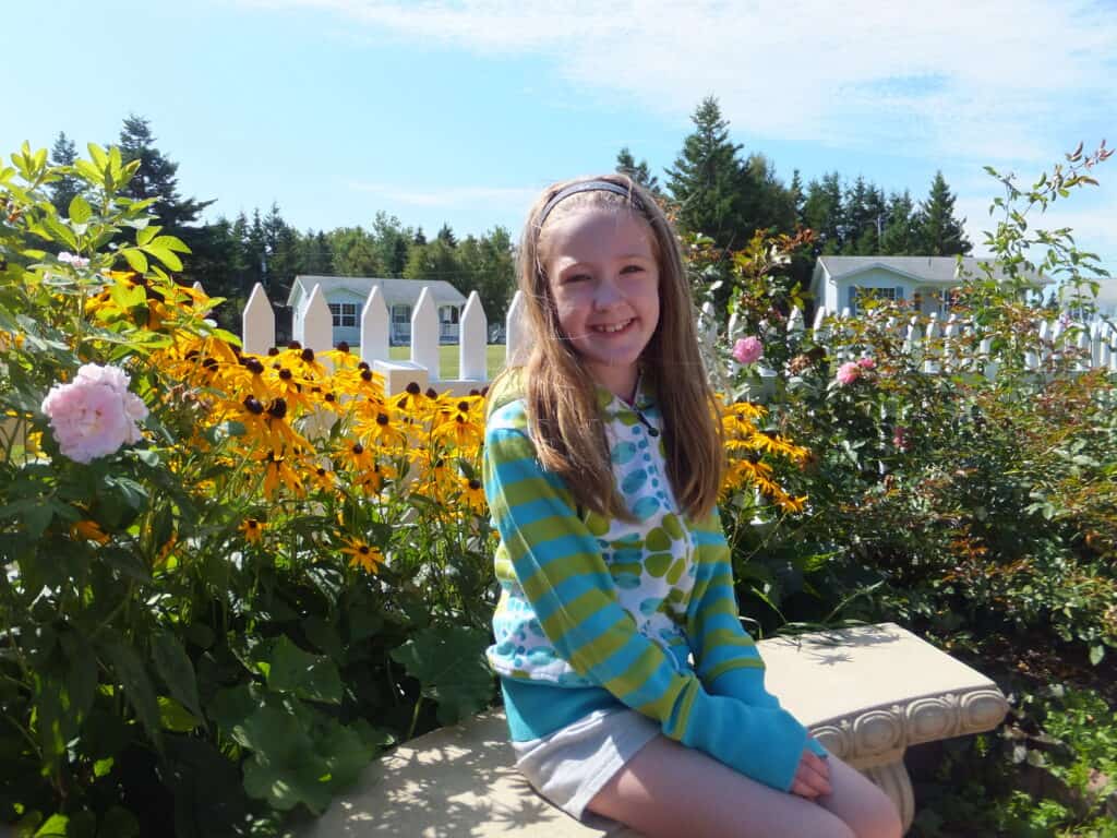 Young girl in blue and green patterned sweatshirt sitting on curved stone garden bench surrounded by pink roses and brown-eyed susan flowers with white picket fence behind and cottages in background.