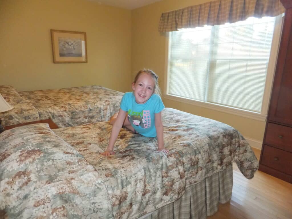 Young girl in bright blue shirt sitting on one of two twin beds with flowered bedding, window in background - Kindred Spirits 3 bedroom cottage.