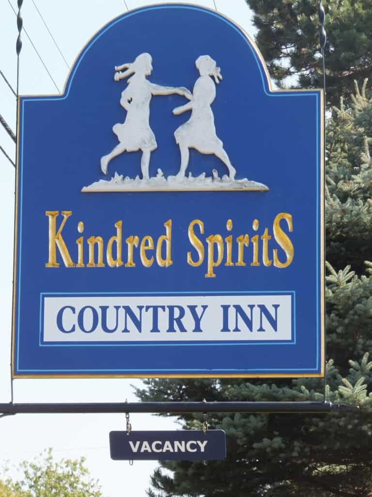 Blue sign with white image of Anne and Diana dancing and text reading Kindred Spirits Country Inn with Vacancy sign hanging on bar under sign.