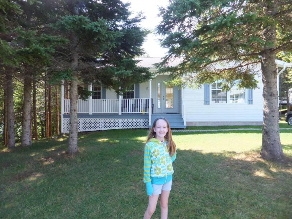 Young girl wearing grey shorts and blue and green patterned sweatshirt standing outside white cottage with grey trim set among trees.