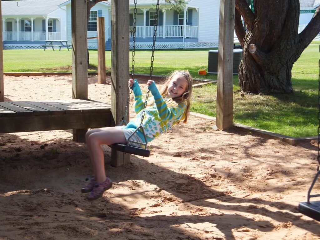 Young girl in blue and green sweater and shorts playing on wooden swing on playground with cottages in background at Kindred Spirits Country Inn & Cottages in Cavendish, Prince Edward Island.