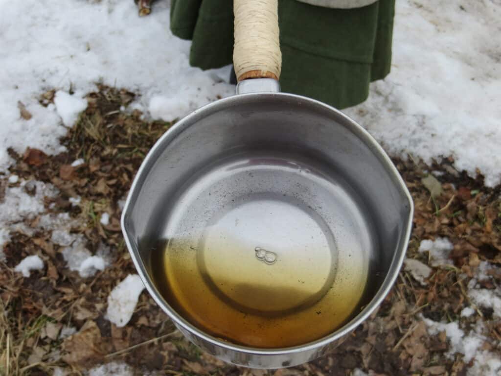 Metal pan with golden coloured liquid held outside above ground covered with plant material and some snow.