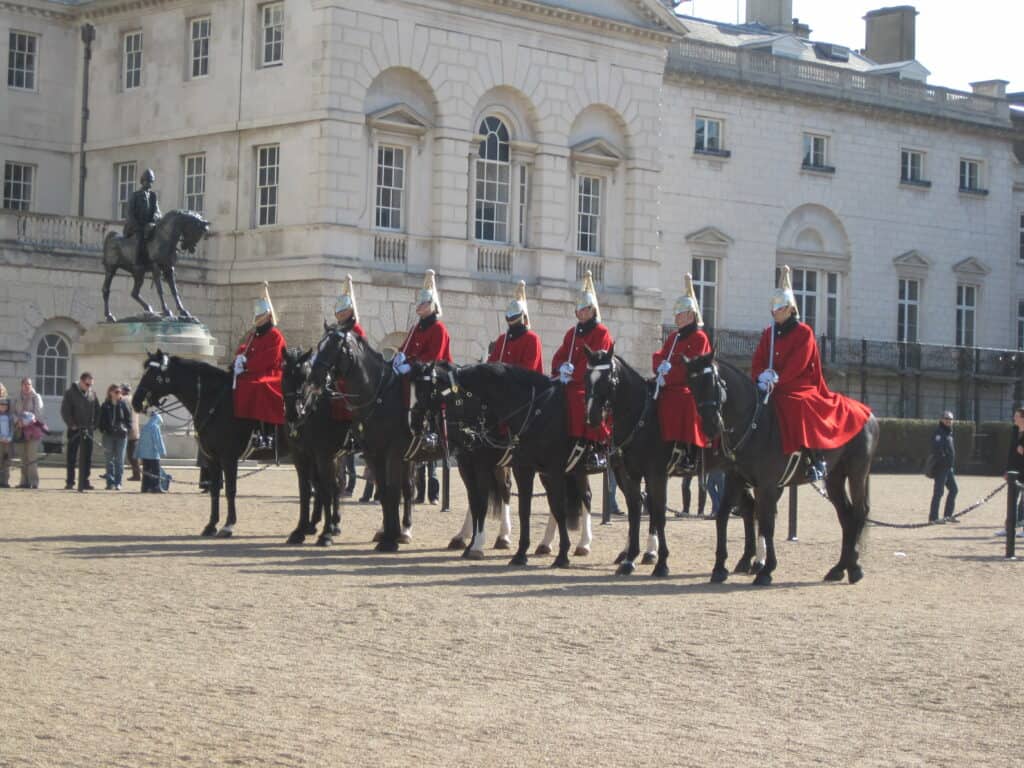 Seven guards in red coats sitting on black horses lined up in front of white stone building in London, England.