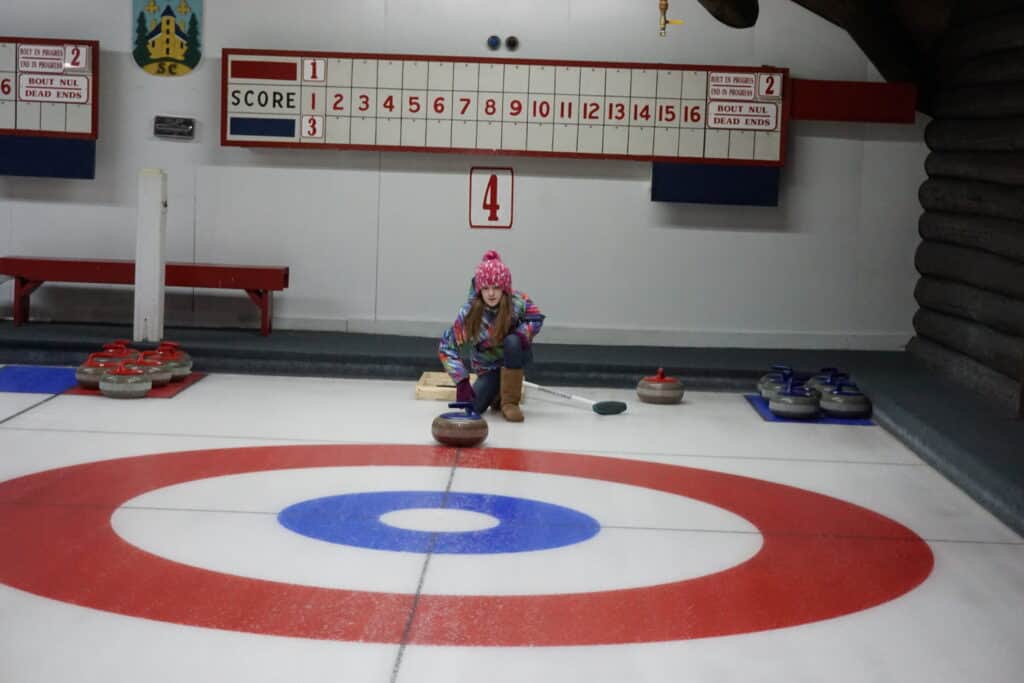 Young girl in multicoloured winter coat and pink hat kneels with curling rock at Chateau Montebello curling rink.